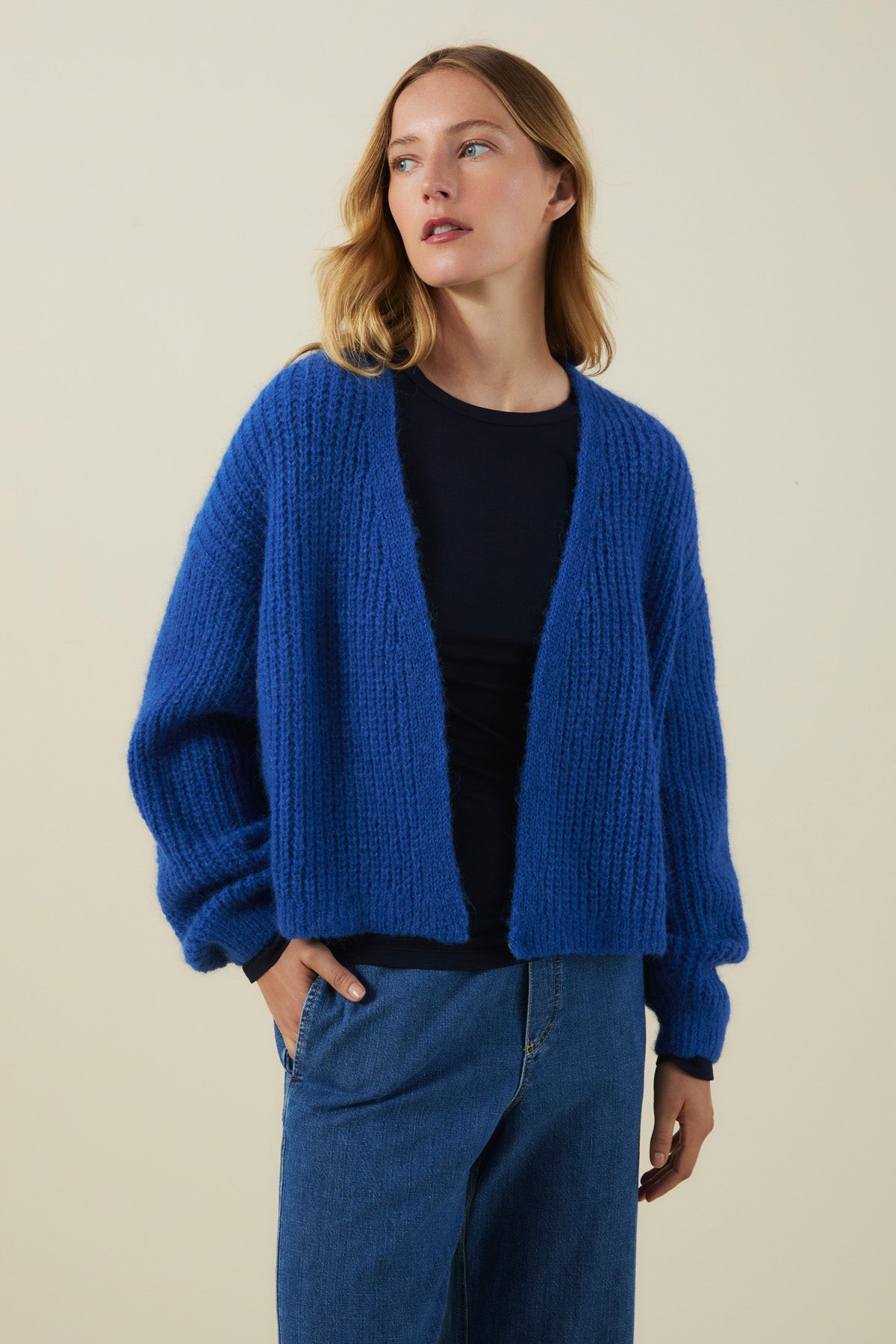 A person models the Velvet by Graham & Spencer RANYA ALPACA CARDIGAN in blue, layered over a black top and blue jeans against a plain beige background, gazing slightly to the side. The chunky alpaca blend knit of the cardigan delivers cozy layering for any occasion.-41976490819777