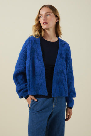 A person models the Velvet by Graham & Spencer RANYA ALPACA CARDIGAN in blue, layered over a black top and blue jeans against a plain beige background, gazing slightly to the side. The chunky alpaca blend knit of the cardigan delivers cozy layering for any occasion.