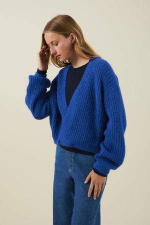A person wearing the Velvet by Graham & Spencer RANYA ALPACA CARDIGAN—a chunky blue alpaca blend layering piece with a relaxed, wrap-style fit—over a navy top and blue jeans, standing against a plain light background, looking down and touching their hair.