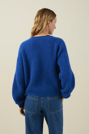 A person with shoulder-length light brown hair models the Velvet by Graham & Spencer RANYA ALPACA CARDIGAN—a chunky blue knit—paired with blue jeans, showcasing cozy layering as they stand with their back to the camera against a neutral background.