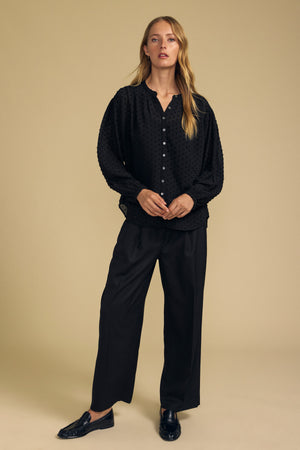 A person wears a black ADRIANA TEXTURED CREPE TOP in textured crepe fabric, paired with loose black trousers and black loafers.