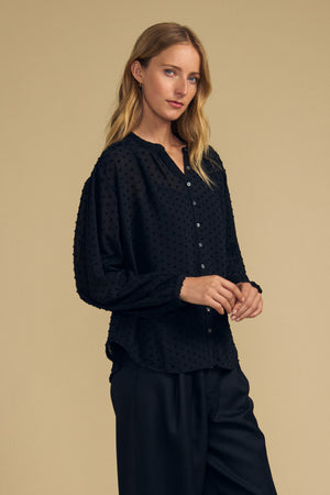 A woman wears a black, long-sleeved ADRIANA TEXTURED CREPE TOP featuring a button-front, textured Swiss dot pattern, and a small collar.