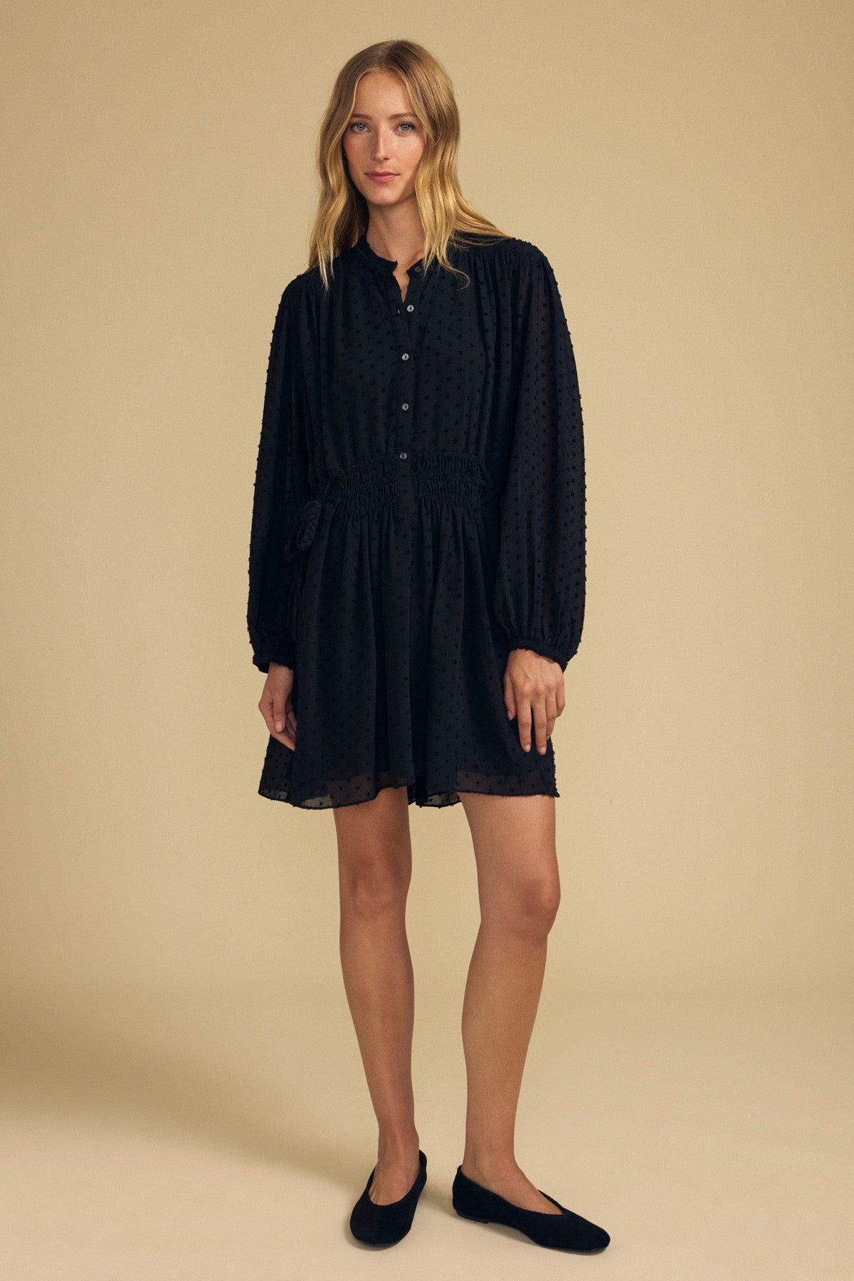 A woman wears a Velvet by Graham & Spencer BRINLEY TEXTURED CREPE DRESS, a black long-sleeved mini dress in textured crepe with a button-down front, gathered elastic waist, and a subtle dotted pattern.-46488137072833