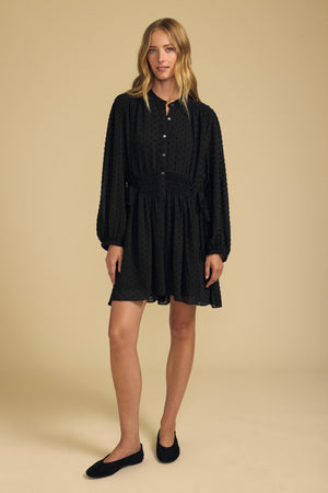 A person wearing a black Velvet by Graham & Spencer BRINLEY TEXTURED CREPE DRESS featuring a button-down front, gathered waist, and long sleeves, paired with black ballet flats.