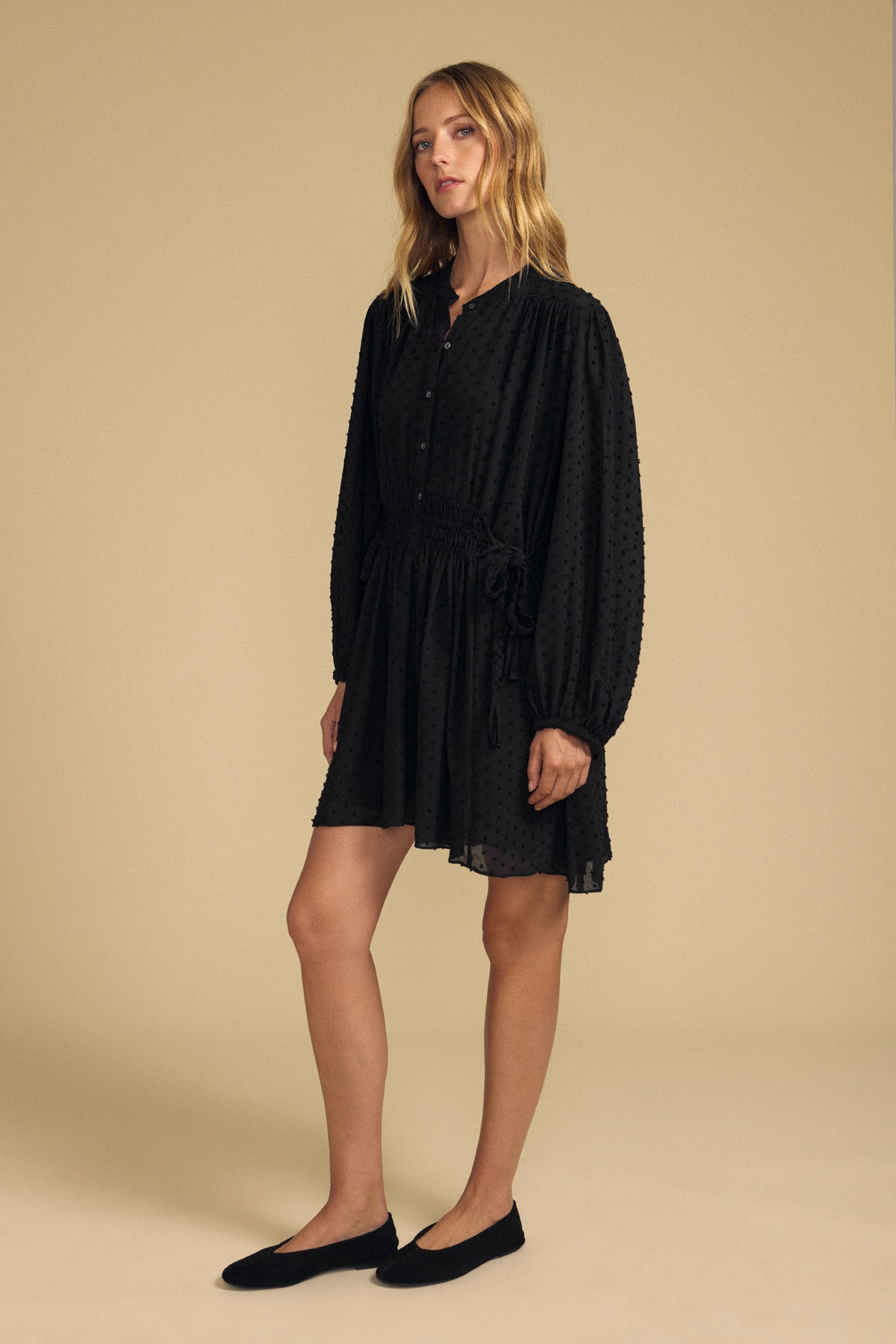 A model wears the Velvet by Graham & Spencer BRINLEY TEXTURED CREPE DRESS, a long-sleeved black button-down mini dress in textured crepe with a drawstring waist, paired with black flats.