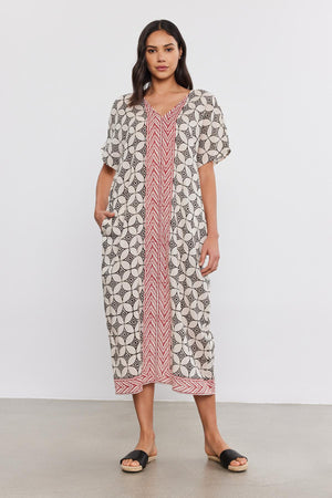 A woman stands facing the camera, wearing an ODESSA KAFTAN DRESS from Velvet by Graham & Spencer with a geometric and stripe pattern, and black sandals on a plain background.