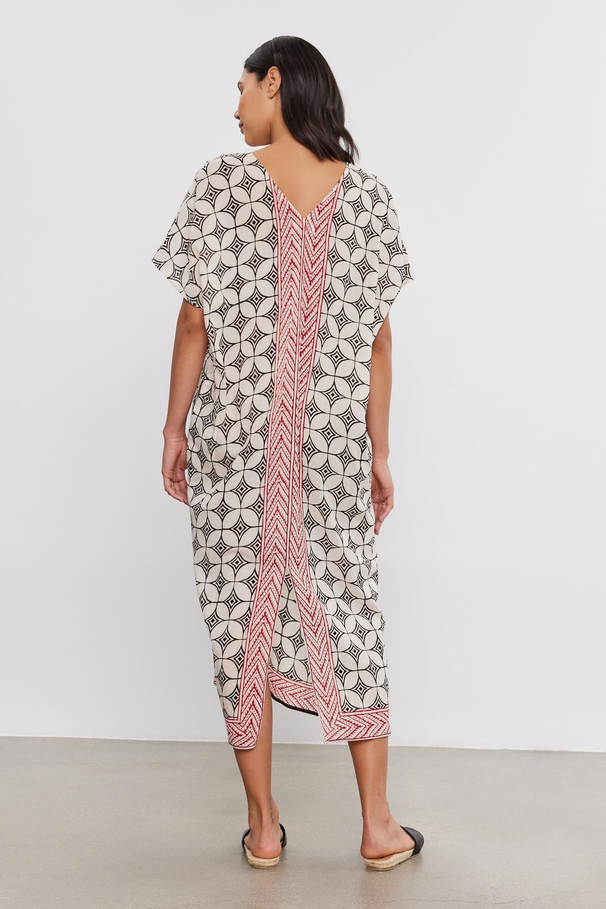 A woman stands with her back to the camera, wearing the ODESSA KAFTAN DRESS by Velvet by Graham & Spencer, a mid-length, lightweight cotton kaftan with a geometric pattern and a bold red stripe down the center.-36910025703617
