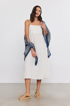 A woman in a white midi dress and patterned blue scarf stands confidently, holding the Velvet by Graham & Spencer sarong wrap with one hand, and wearing gold sandals.