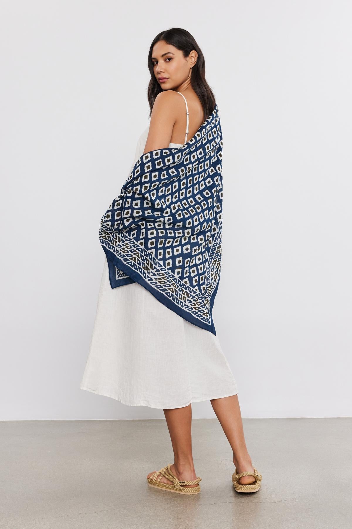 A woman in a white dress and espadrilles, draped in a Velvet by Graham & Spencer SARONG WRAP, stands looking over her shoulder against a plain background.-36752950591681