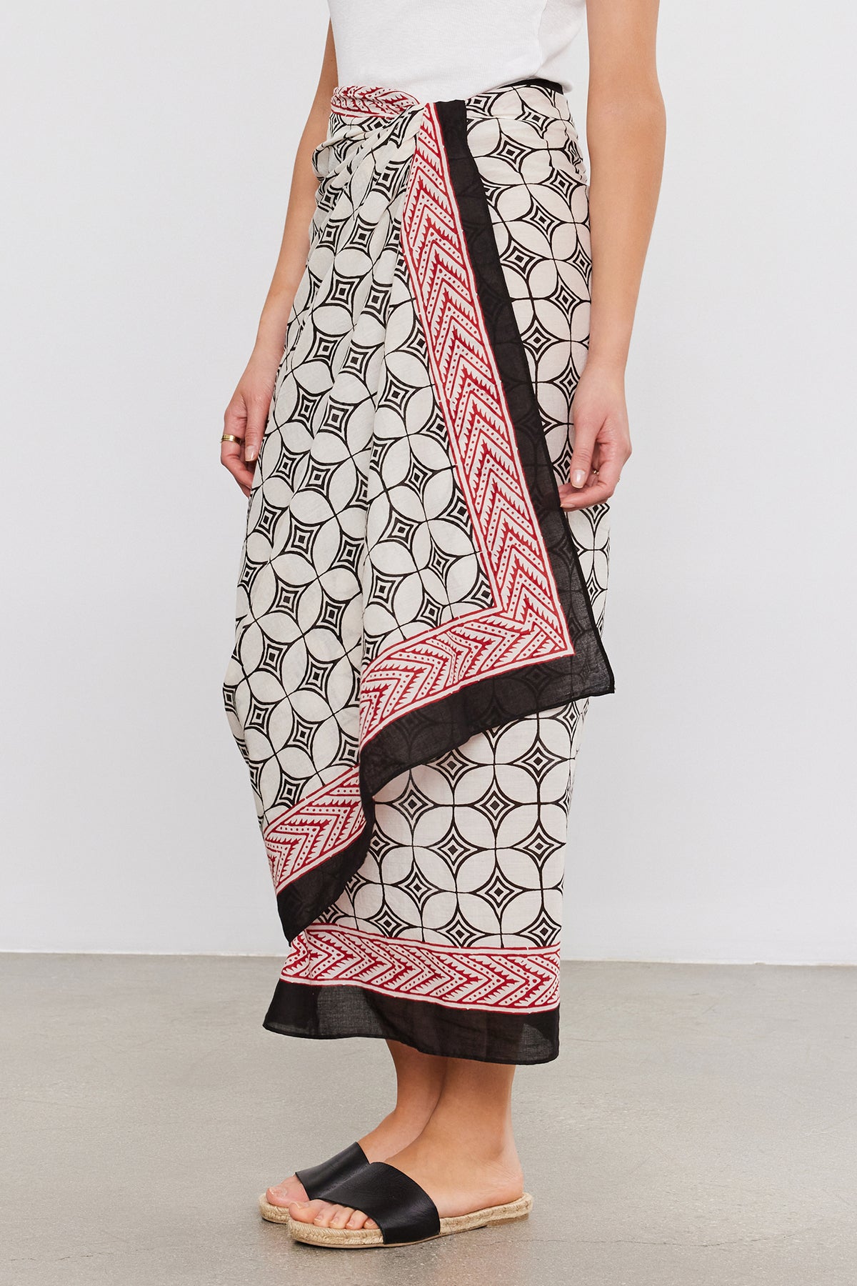 Person wearing a Velvet by Graham & Spencer SARONG WRAP with black and red borders, reminiscent of a sarong, stands on a gray floor, paired with a white top and black slide sandals.-37367009575105