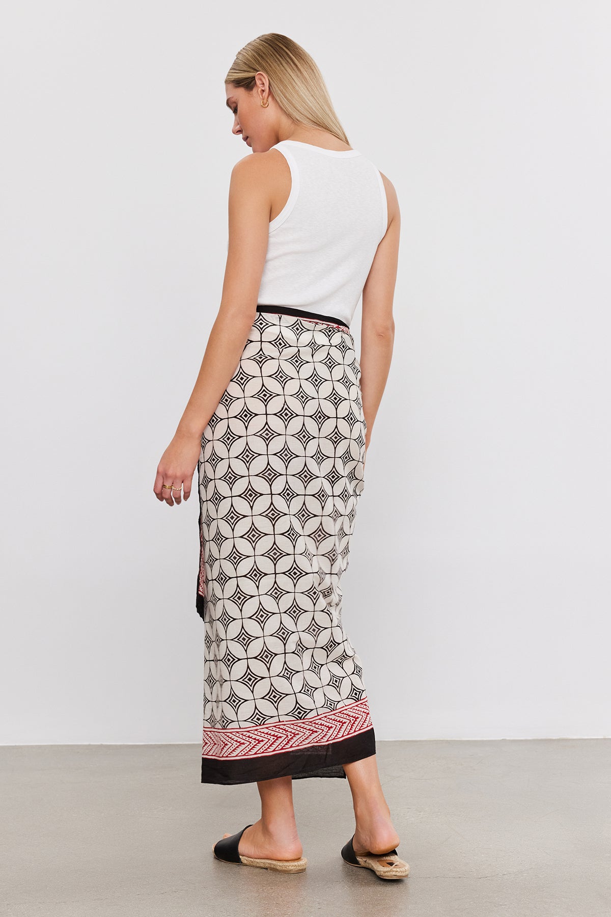 A person with long blonde hair, wearing a white sleeveless top and a Velvet by Graham & Spencer SARONG WRAP featuring vibrant prints, stands with their back towards the camera in a minimalist indoor setting.-37367009607873