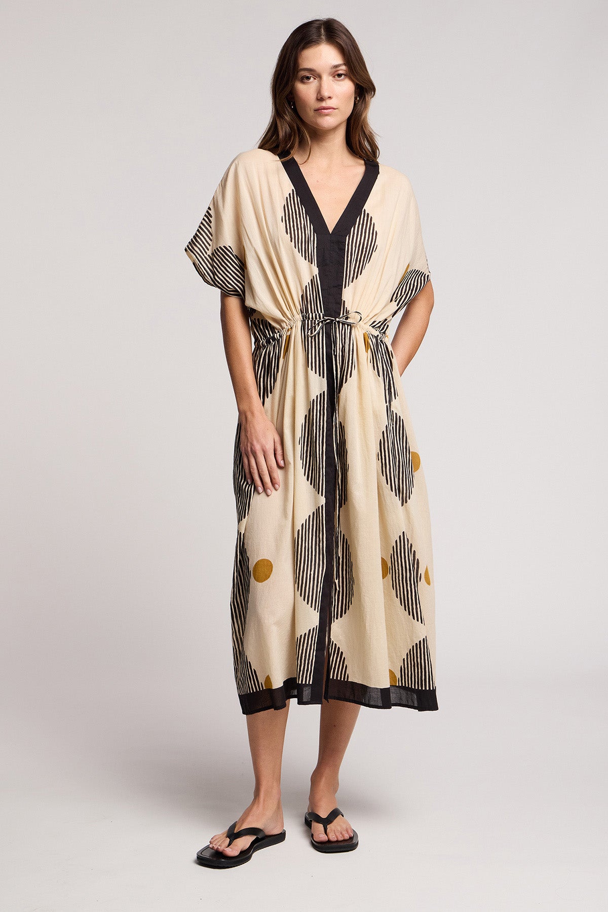 A woman stands against a plain background in the AMIRA DRESS by Velvet by Graham & Spencer, a beige midi dress with black stripes, yellow dots, black trim, and a tie waist, styled with flip-flops.-41370929823937