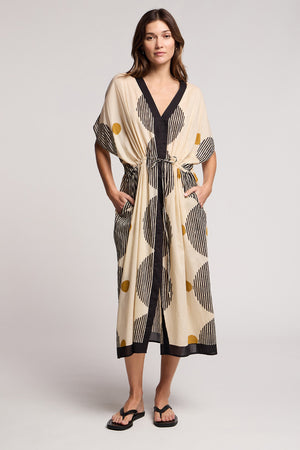 A woman wears the AMIRA DRESS by Velvet by Graham & Spencer—a travel-ready beige kimono-style dress with black stripes, yellow dots, short sleeves, tie waist, pockets—and pairs it with black sandals.