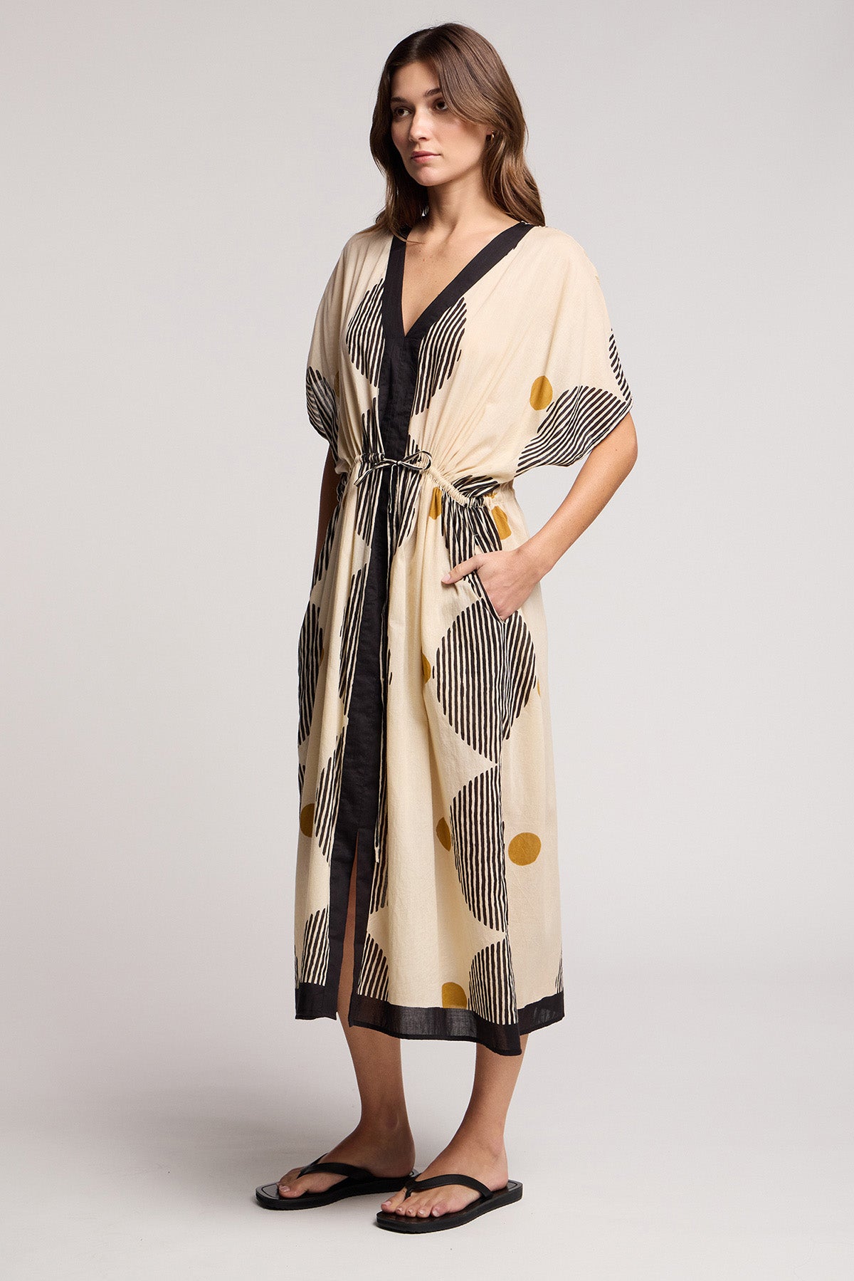 A woman stands against a plain background wearing the AMIRA DRESS by Velvet by Graham & Spencer—a beige, black, and yellow patterned travel-ready dress with a belt—paired with black sandals.-41370929692865