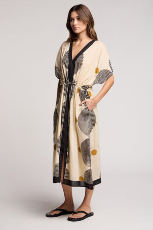 A woman stands against a plain background wearing the AMIRA DRESS by Velvet by Graham & Spencer—a beige, black, and yellow patterned travel-ready dress with a belt—paired with black sandals.