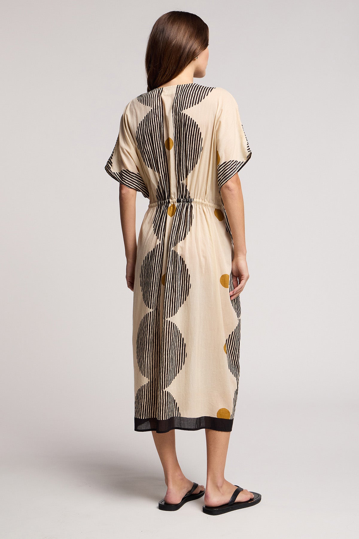 A woman stands facing away, wearing the AMIRA DRESS by Velvet by Graham & Spencer—a beige cotton kaftan with black stripes and bold black and yellow dots, paired with black sandals. A travel-ready look for any occasion.-41370929660097