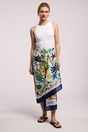 A woman stands against a plain background wearing a white sleeveless top, brown flip-flops, and the ROBI SARONG by Velvet by Graham & Spencer—a vibrant cotton wrap skirt with botanical and marine motifs.