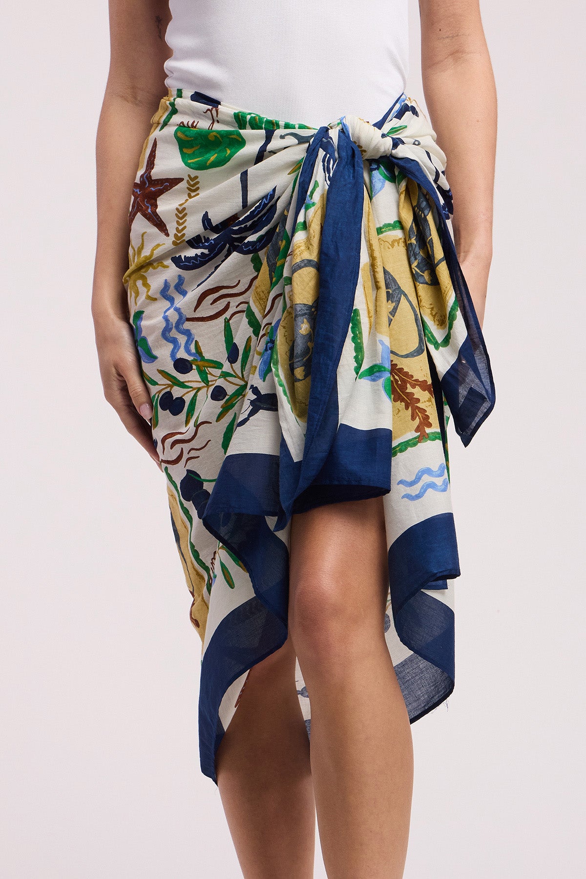 A person wears the ROBI SARONG by Velvet by Graham & Spencer—a vibrant printed cotton sarong with a navy blue border, styled at the waist over a white top, perfect as a chic beach cover-up.