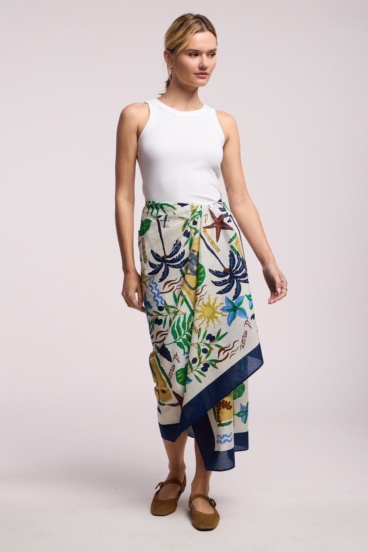 A woman wearing a white sleeveless top, brown shoes, and the ROBI SARONG by Velvet by Graham & Spencer—featuring a colorful tropical-patterned cotton wrap skirt—stands against a plain background.