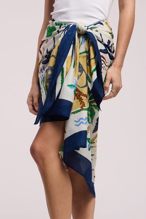 A person wears Velvet by Graham & Spencer's ROBI SARONG, a printed cotton cover-up with a navy border, tied at the waist over a white tank top for a stylish beach look.