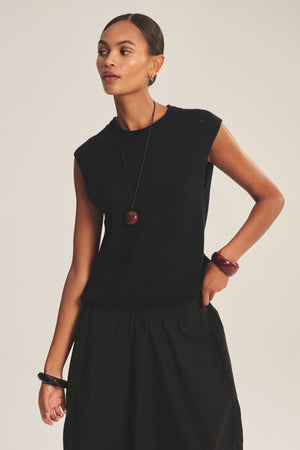A woman wears the VANNY SWEATER by Velvet by Graham & Spencer—a sleeveless black top with subtle pointelle detail—paired with a black skirt, long necklace, and chunky bracelets for an effortlessly chic transitional-weather look.
