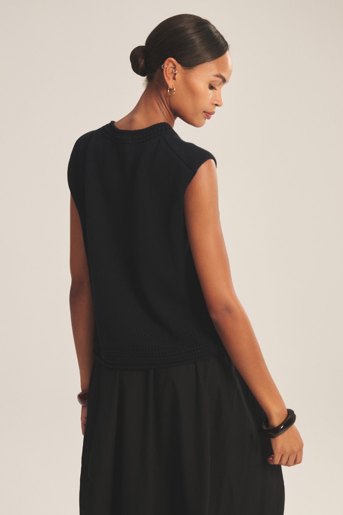 A woman with dark hair in a low bun wears the VANNY SWEATER by Velvet by Graham & Spencer, a sleeveless black top with pointelle detailing, and a skirt, standing with her back to the camera against a neutral background.-41338487242945