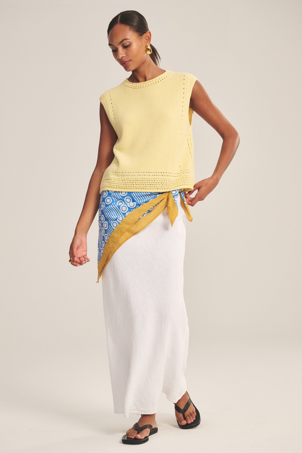 A woman styles the VANNY SWEATER by Velvet by Graham & Spencer—a sleeveless yellow pima cotton vest—with a white maxi skirt, blue and yellow scarf at the waist, black sandals, and gold hoops for a chic transitional-weather look.