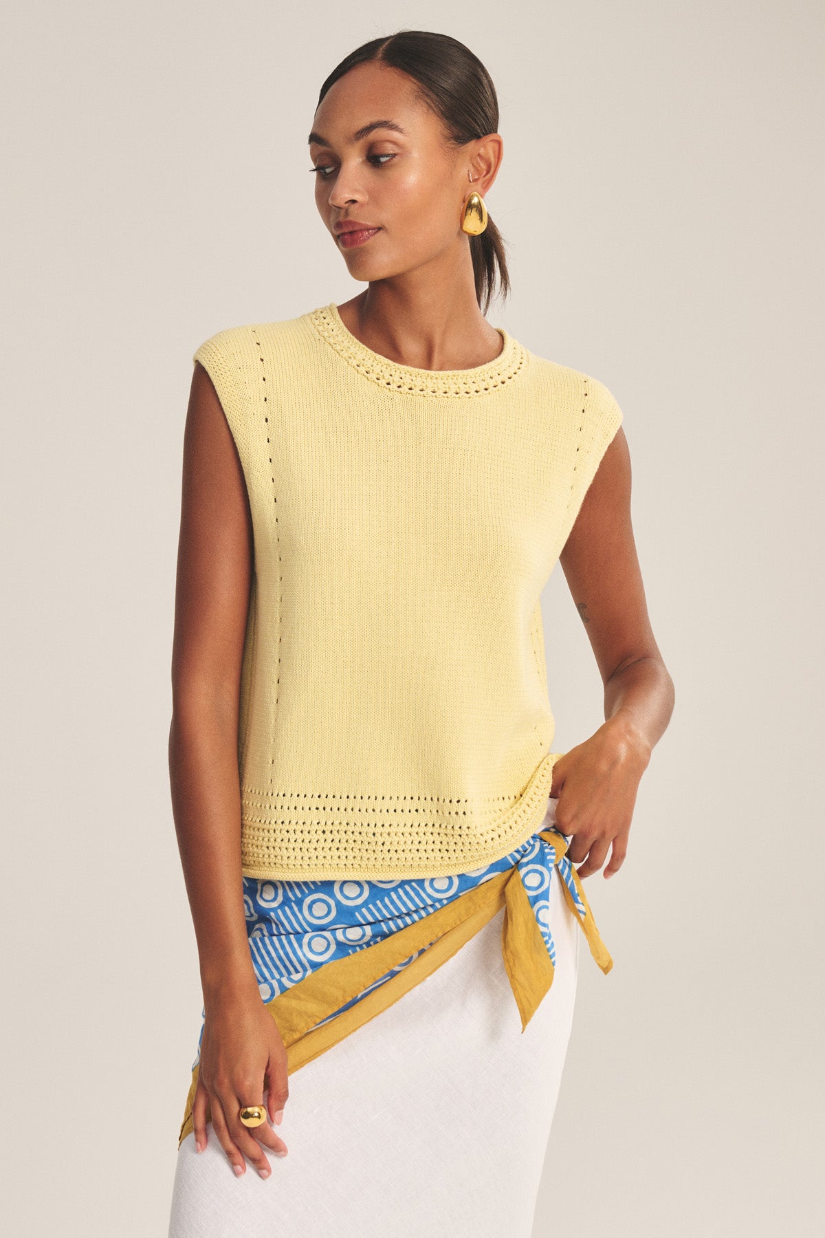 A woman wears the Velvet by Graham & Spencer VANNY SWEATER—a sleeveless yellow pima cotton vest with pointelle details—styled with gold hoop earrings and a blue-and-white wrap skirt with yellow accents; ideal for transitional weather.-41128868151489