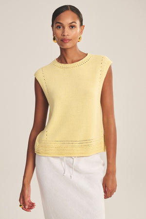 A woman wears the VANNY SWEATER by Velvet by Graham & Spencer—a sleeveless yellow pointelle vest—paired with a white skirt against a plain background, ideal for transitional weather.