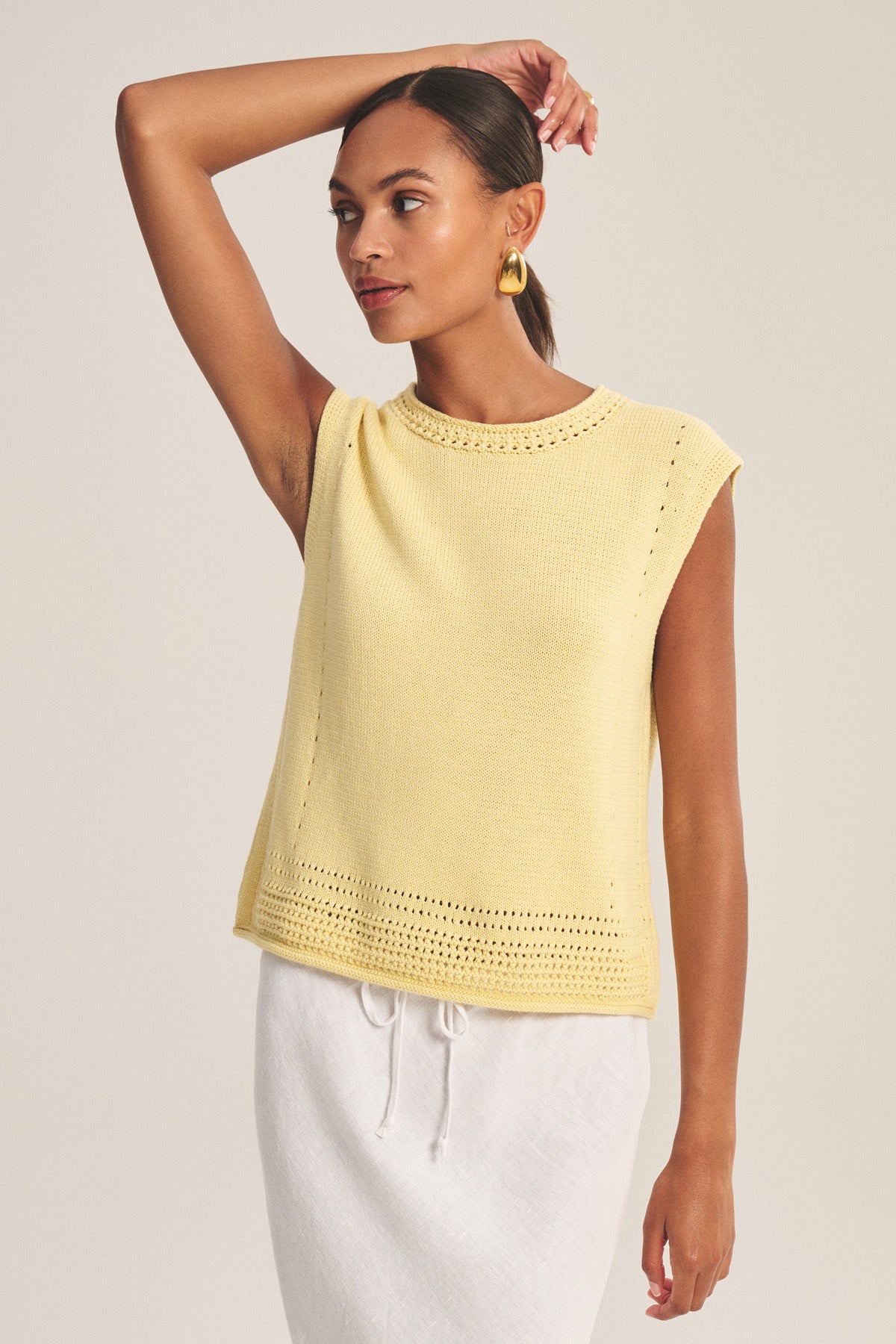 A woman in the VANNY SWEATER by Velvet by Graham & Spencer—a sleeveless yellow pointelle vest—pairs it with a white drawstring skirt, striking a pose against a neutral background for chic transitional-weather style.