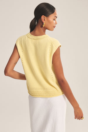 A woman with dark hair in a low ponytail wears the Velvet by Graham & Spencer VANNY SWEATER—a sleeveless yellow pima cotton vest—paired with a white skirt, standing with her back to the camera against a plain background.