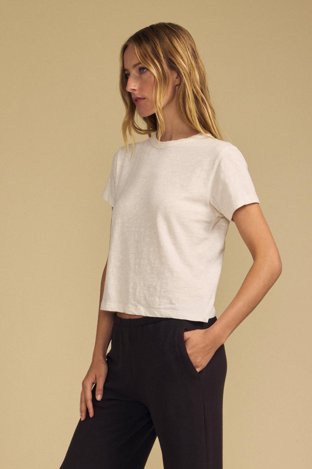 A woman stands against a beige background, wearing the relaxed fit LEIGH TEE in white—crafted from cotton jersey by Velvet by Graham & Spencer—and black pants, with one hand in her pocket as she looks to the side.-41976735367361