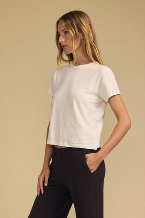 A woman stands against a beige background, wearing the relaxed fit LEIGH TEE in white—crafted from cotton jersey by Velvet by Graham & Spencer—and black pants, with one hand in her pocket as she looks to the side.