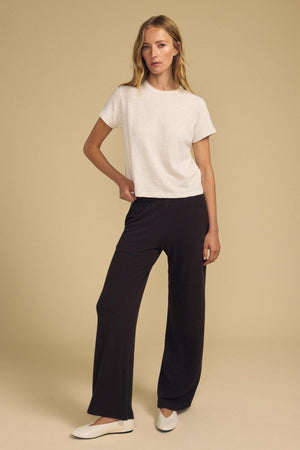 A person stands against a neutral background wearing the LEIGH TEE by Velvet by Graham & Spencer—a white, relaxed fit cotton jersey short-sleeve t-shirt—paired with black wide-leg pants and white flats.