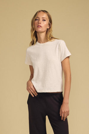 A person with long blonde hair wears the LEIGH TEE by Velvet by Graham & Spencer—a vintage-inspired tee crafted from plain white cotton jersey—paired with dark pants, standing against a neutral beige background.