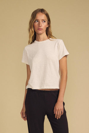A person stands against a beige background, wearing the LEIGH TEE by Velvet by Graham & Spencer—a light beige cotton jersey short-sleeve t-shirt with a relaxed fit—paired with dark pants.