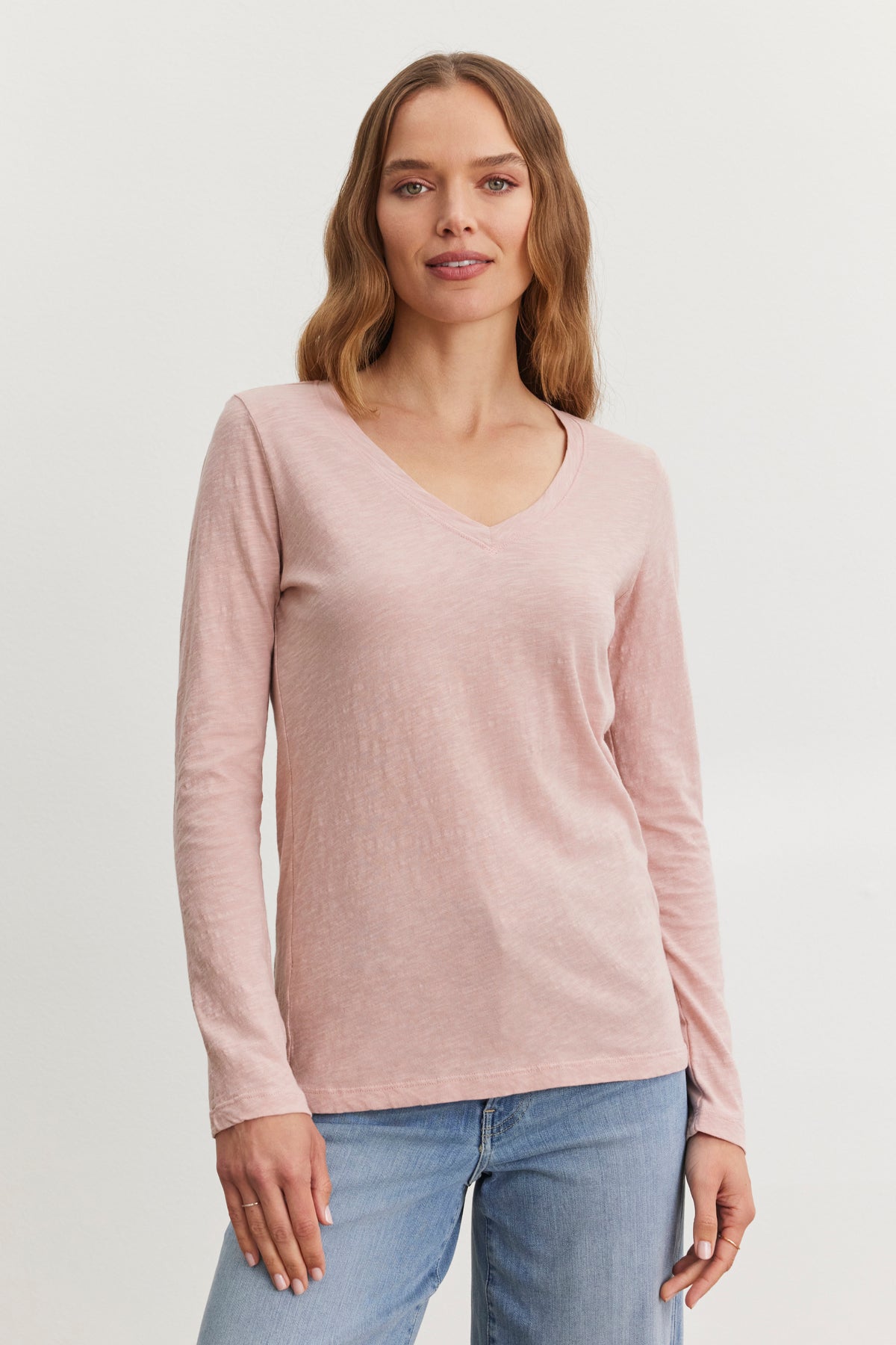   A person wearing the BLAIRE TEE, a light pink textured cotton long-sleeve V-neck shirt from Velvet by Graham & Spencer, and blue jeans poses against a plain background. 