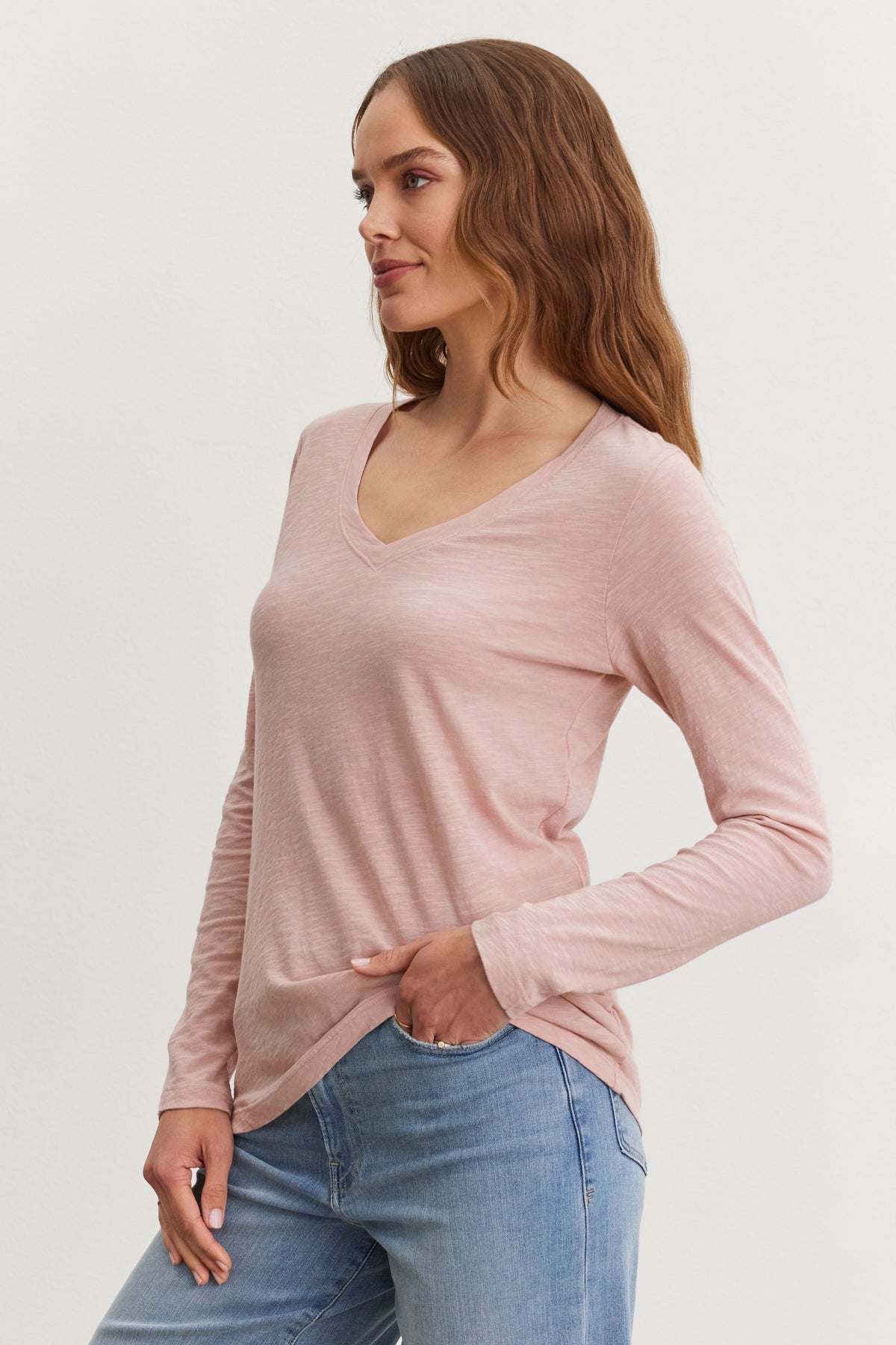   A person wearing a Blaire Tee by Velvet by Graham & Spencer, which is a light pink, v-neckline long-sleeve shirt made of textured cotton, and blue jeans stands against a plain background, looking to the side. 