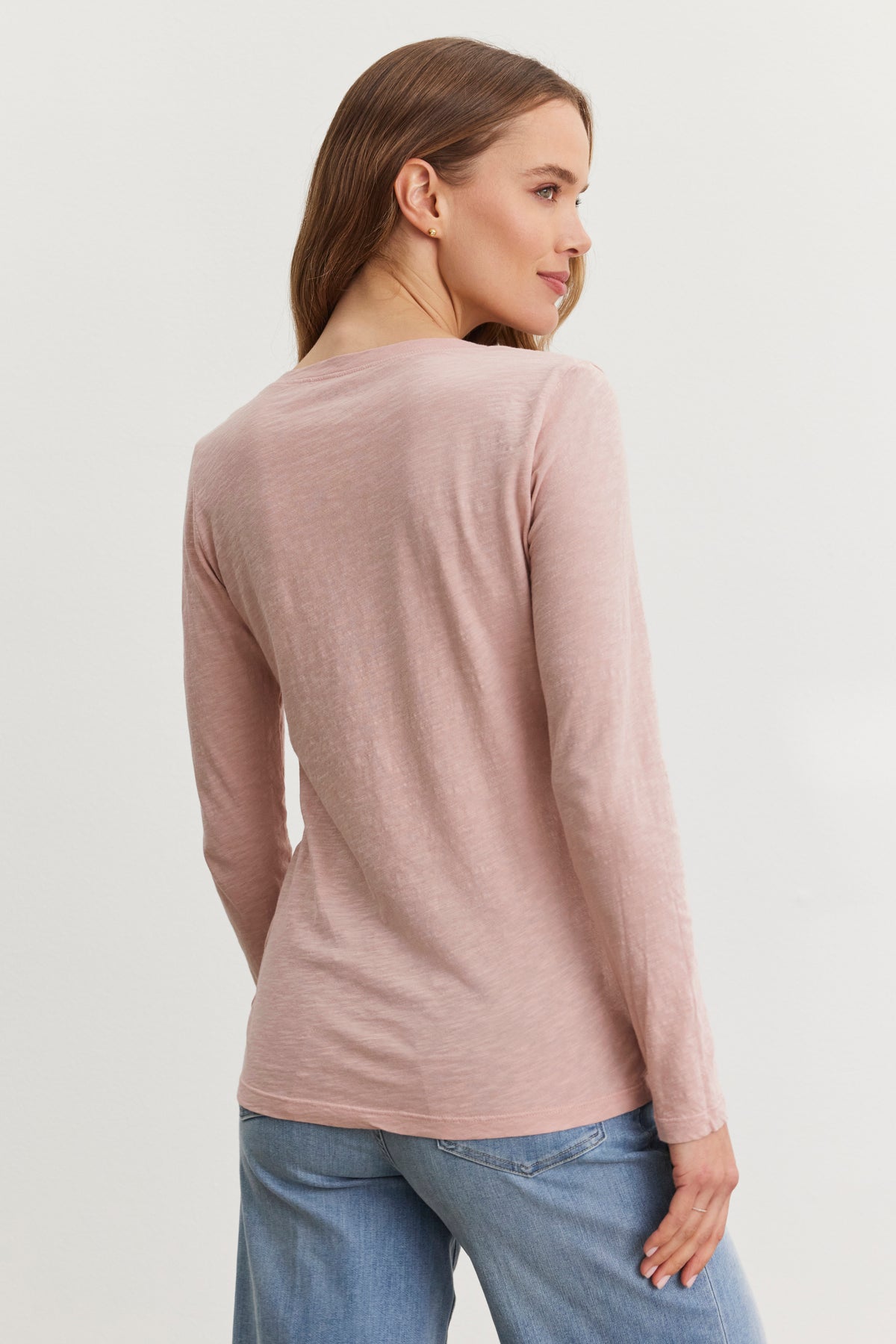   A woman wearing the Velvet by Graham & Spencer BLAIRE TEE, a pink textured cotton long-sleeve shirt, paired with blue jeans, is shown from the side against a light background. 