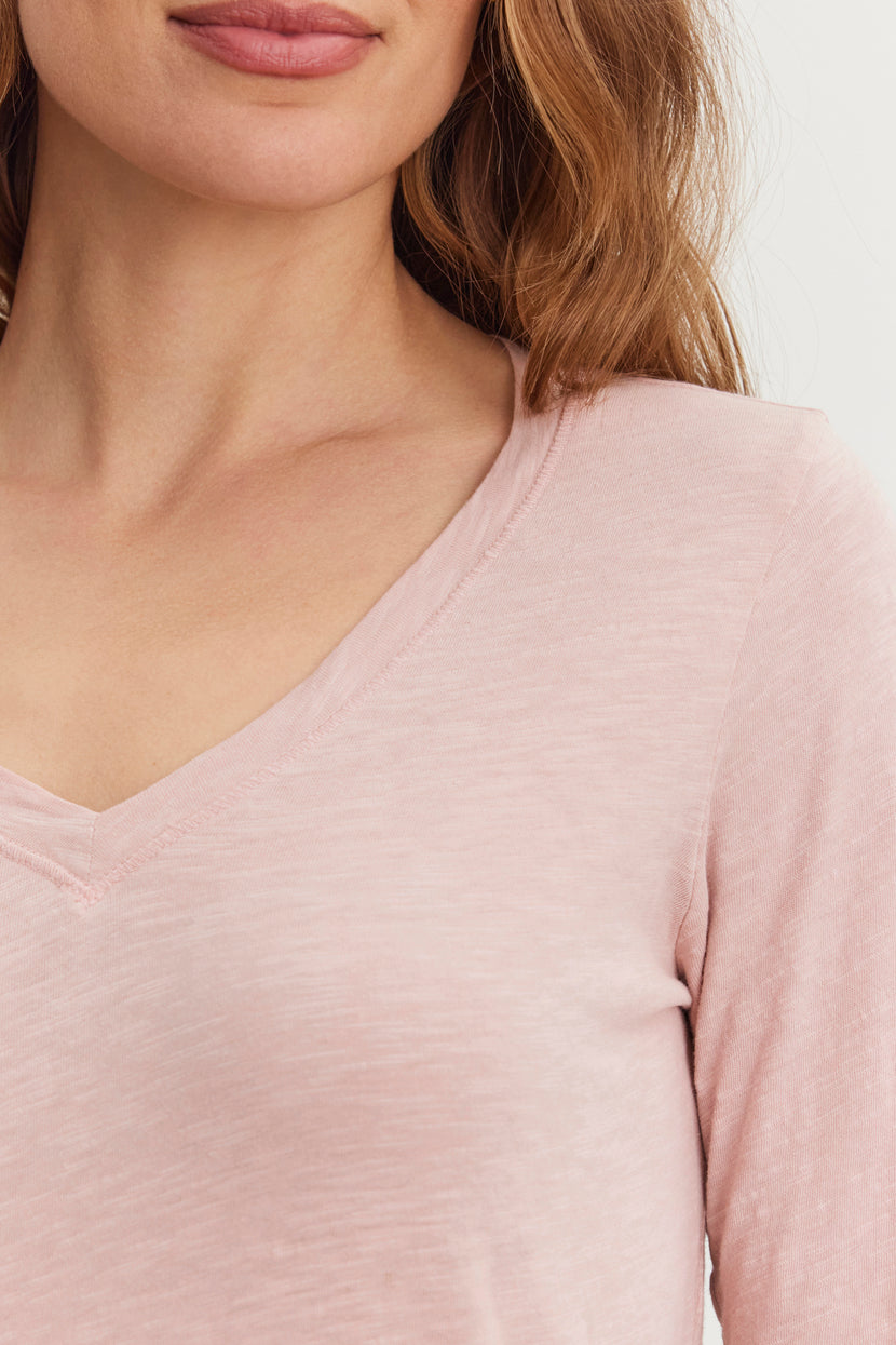 Close-up of a woman wearing the light pink BLAIRE TEE from Velvet by Graham & Spencer, featuring a V-neck design with textured cotton, highlighting her neck and shoulders against a plain background.