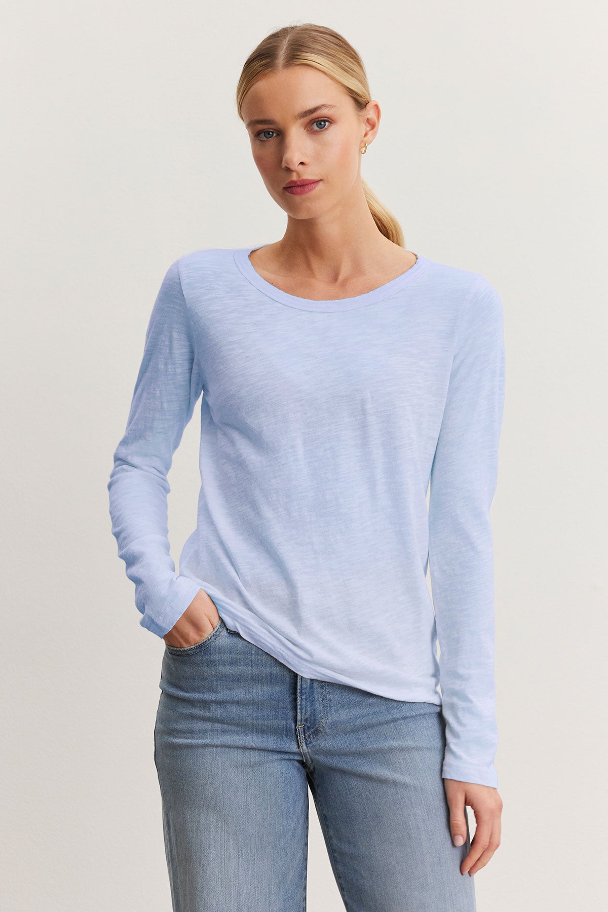   A person with long hair in a ponytail, wearing a Velvet by Graham & Spencer LIZZIE TEE—light blue with a classic crew neckline—and jeans, stands against a plain background. 