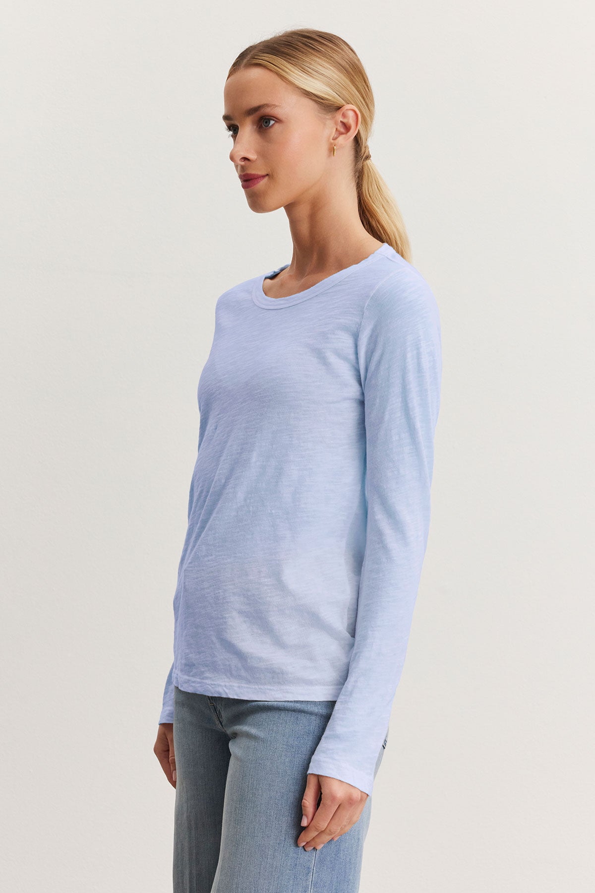   A person models the Velvet by Graham & Spencer LIZZIE TEE, featuring a light blue textured cotton slub with a classic crew neckline, paired with jeans against a plain backdrop. 