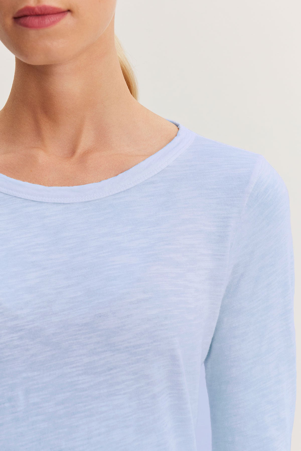   Close-up of a person wearing the LIZZIE TEE by Velvet by Graham & Spencer, a light blue long-sleeve with a classic crew neckline, facing forward. The textured cotton slub enriches the neutral backdrop. 