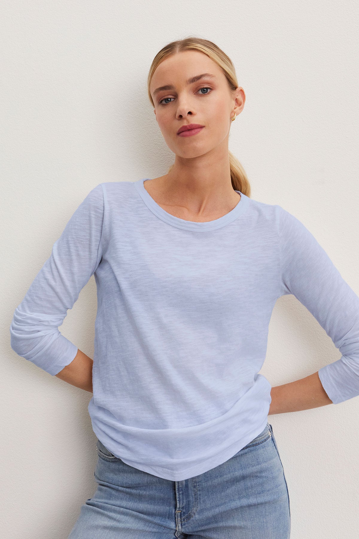   A person in a Velvet by Graham & Spencer LIZZIE TEE, featuring a classic crew neck and long sleeves in light blue, pairs it with jeans while standing against a plain background. 