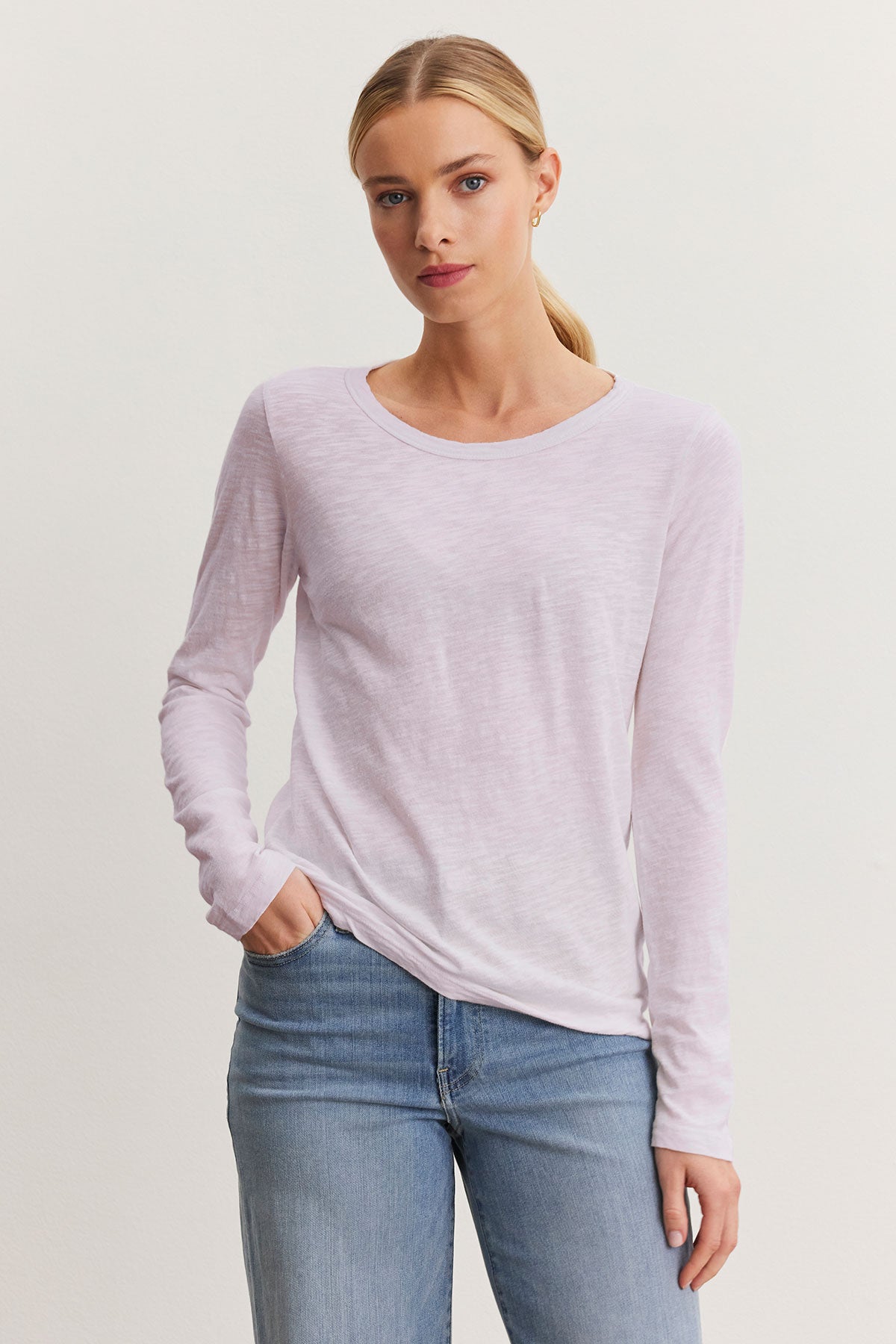   A person wearing the LIZZIE TEE, a light pink textured cotton slub long-sleeve shirt from Velvet by Graham & Spencer, pairs it with blue jeans while standing against a plain background, hand in pocket. 