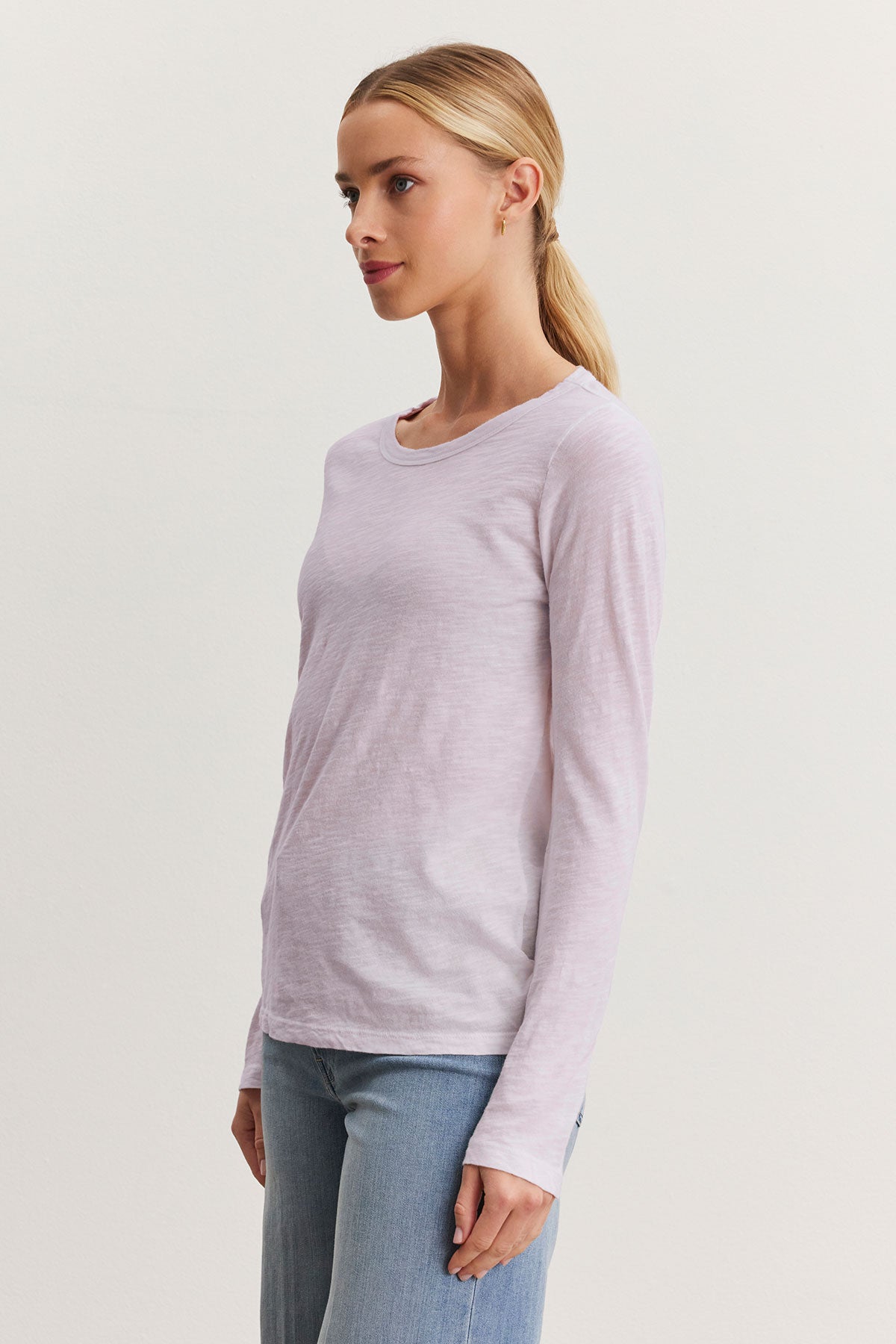   A person with long hair in a ponytail, wearing the LIZZIE TEE from Velvet by Graham & Spencer—a light pink, long-sleeve shirt with a classic crew neckline—and blue jeans, stands sideways against a plain background. 