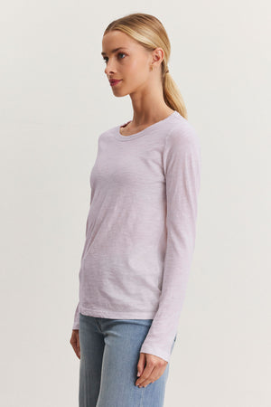 A person with long hair in a ponytail, wearing the LIZZIE TEE from Velvet by Graham & Spencer—a light pink, long-sleeve shirt with a classic crew neckline—and blue jeans, stands sideways against a plain background.