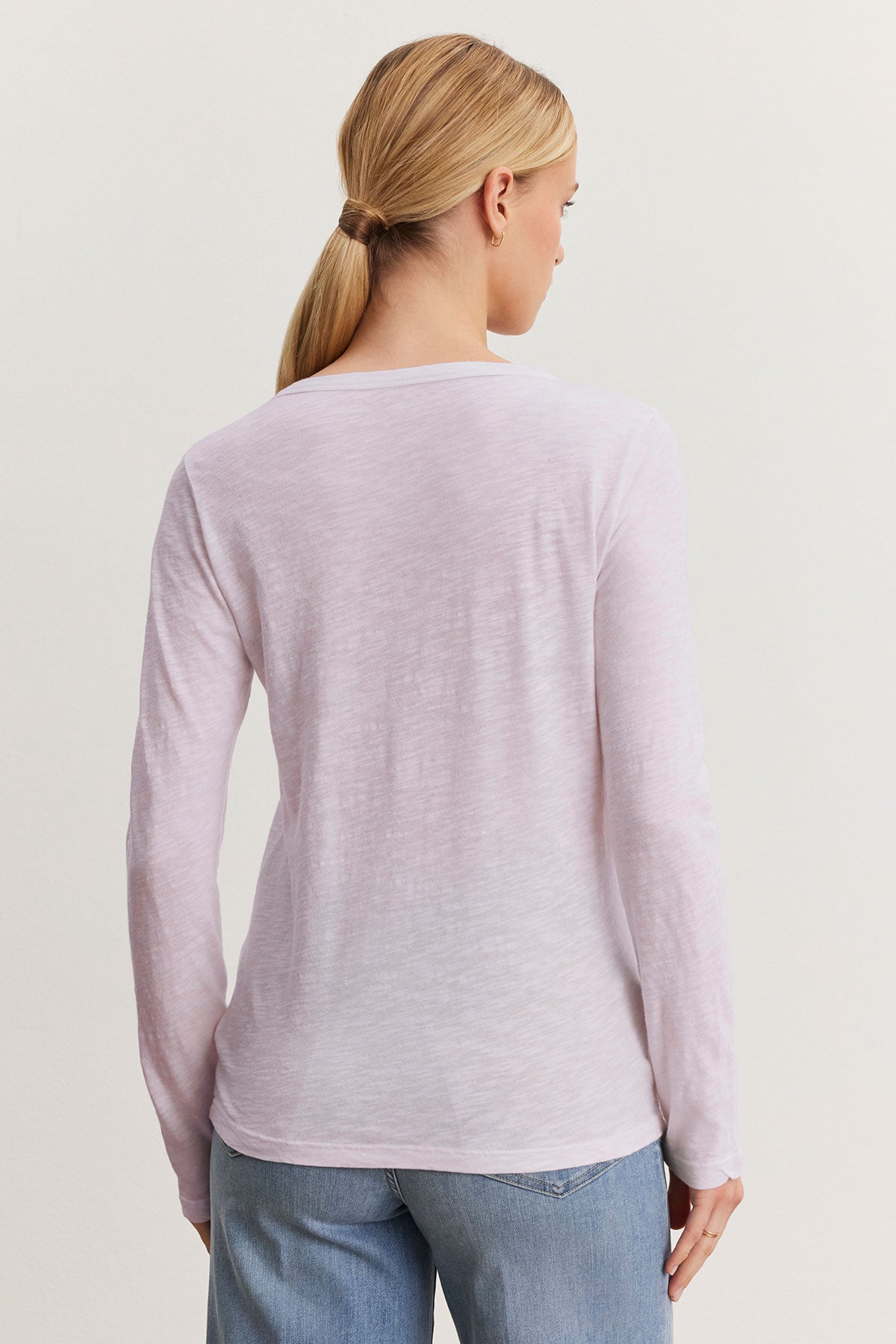   A person with blonde hair in a ponytail is wearing the LIZZIE TEE by Velvet by Graham & Spencer, a light pink long-sleeve shirt with a classic crew neckline, paired with blue jeans and facing away. 