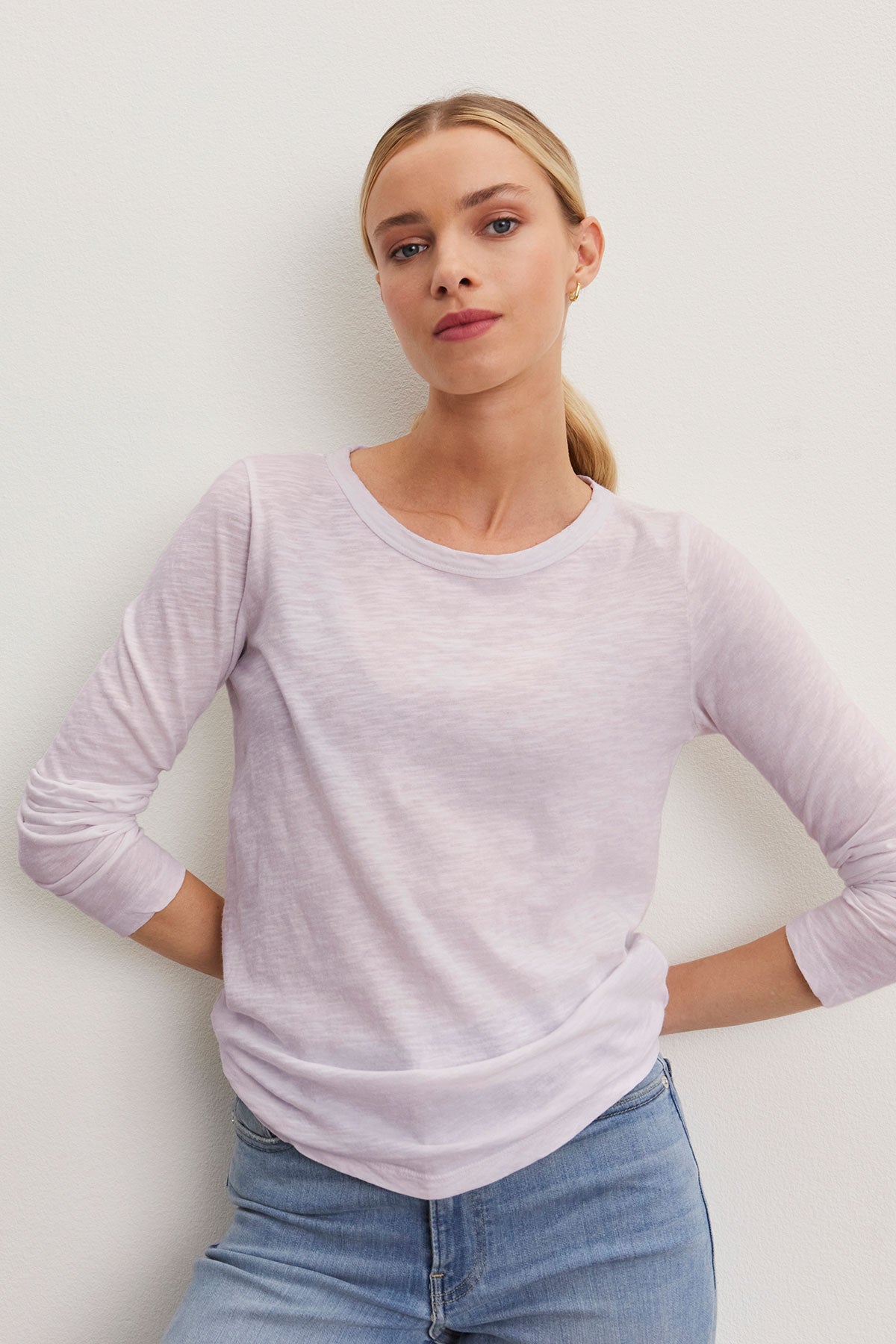 A person stands against a plain background, wearing the LIZZIE TEE from Velvet by Graham & Spencer—a light purple long-sleeve top with a classic crew neckline—paired with casual blue jeans.-38892051333313