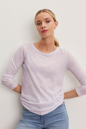 A person stands against a plain background, wearing the LIZZIE TEE from Velvet by Graham & Spencer—a light purple long-sleeve top with a classic crew neckline—paired with casual blue jeans.